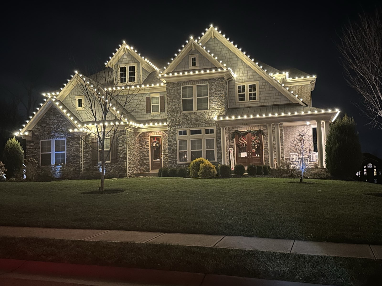 Two-story stone home with warm white C9 LED Christmas lights along the roofline and porch - professional installation